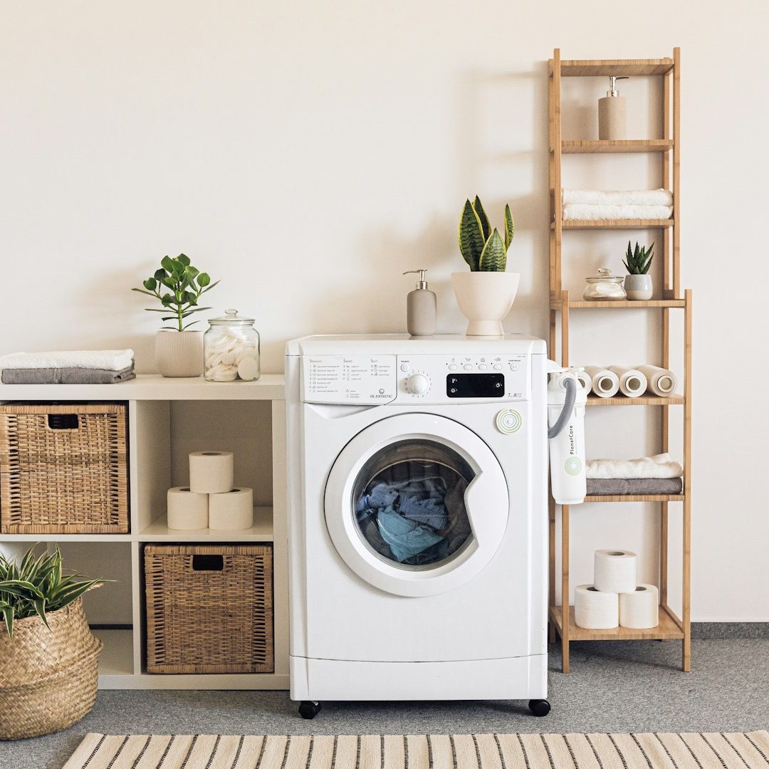 Moderne Waschmaschine neben Regalen mit Pflanzen und Aufbewahrungskörben in heller Umgebung.
